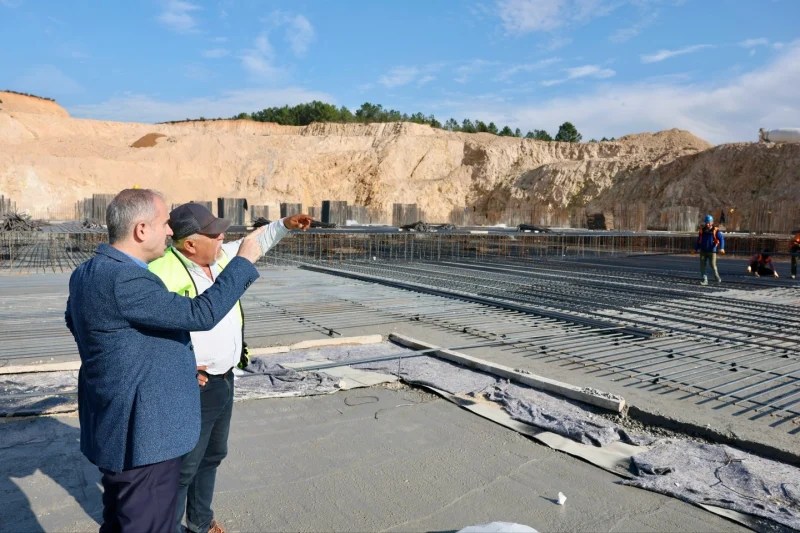 Çayırova Belediye Başkanı Bünyamin Çiftçi, inşaat hâlindeki Çayırova Devlet Hastanesi'nin şantiye alanında yer alan temel beton serim çalışmalarını gözlemliyor. Arka planda inşaat alanı ve işçiler yer alıyor.