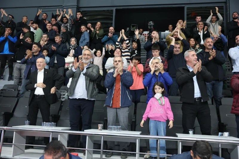 Bir basketbol maçında taraftarlar, ellerini çırparak ve coşkuyla takımlarını destekliyor. Çeşitli yaşlardan ve cinsiyetlerden insanlar, sahneyi izleyen kalabalık içinde yer alıyor.