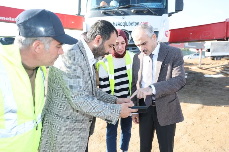 Çayırova Belediye Başkanı Bünyamin Çiftçi, yeni meslek lisesi inşaatında inceleme yaparken, yanında inşaat çalışanları ve bir kadın yer alıyor. Başkan, tablette bilgi alırken görünmektedir.