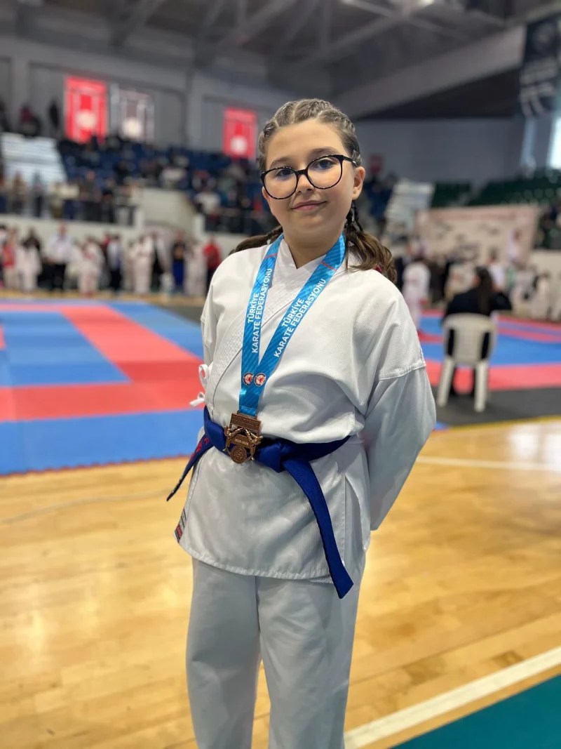 11 yaşındaki karate sporcusu Hayal Nur Kurt, mavi kemerle birlikte bronz madalya ile poz veriyor. Arka planda karate müsabakaları devam ediyor.