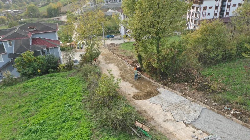 Bahçecik Kılıçarslan Mahallesi'nde sokak yenileme çalışmaları, ağaçların yanında bir işçi tarafından bir parke yolun döşenmesiyle devam ediyor. Arka planda, yerleşim alanları ve yeşil alanlar görünmekte.