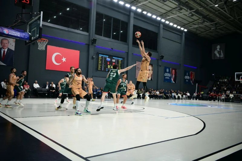 Basketbol maçı sırasında bir oyuncunun şut atma anı, arka planda Türk bayrağı ve bazı izleyiciler görünmekte.