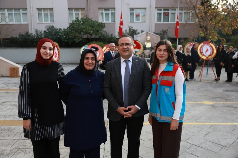 Dört kişi, anma etkinliği sırasında bir arada fotoğraf çektiriyor. Arka planda Türk bayrakları ve çelenkler yer alıyor.