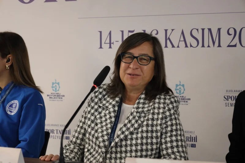 Bir sempozyumda konuşan kadın akademisyen, Kocaeli Spor Tarihi Sempozyumu için hazırlanan masanın önünde gülümsemekte. Arkada başka katılımcılar yer almakta.