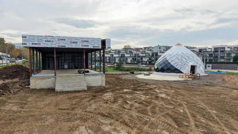 Başiskele'de inşaat halindeki Gün Evi ve Kadın El Emeği Mağazası, modern mimarisiyle dikkat çekiyor. Sağda yer alan çatı formu ve soldaki yapı, kadınlara yönelik sosyal projeleri simgeliyor. Çevre düzenlemesi ve inşaat süreci görünmekte.