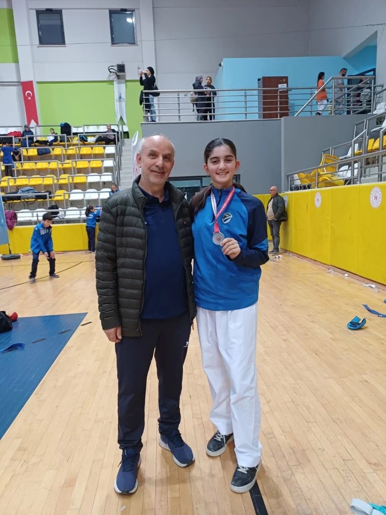 Kartepe Belediyespor karate takımı sporcusu, madalya ile birlikte antrenörüyle birlikte poz veriyor, arka planda spor salonu görünmekte.