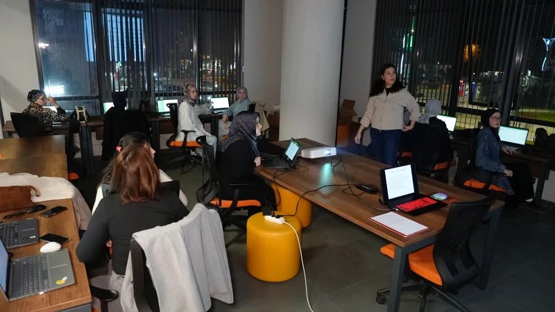 Bir eğitim salonunda bilgisayarlarla çalışan kursiyerlerin olduğu görüntü. Arkada bir eğitmen sunum yapıyor.