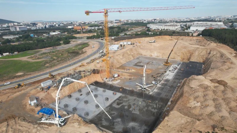 Çayırova Devlet Hastanesi inşaat alanında temel beton dökülüyor. İnşaatta beton pompaları ve vinçler kullanılıyor. Geniş bir inşaat alanı görünümünde, çevresinde inşaat ekipmanları ve araçlar yer alıyor.
