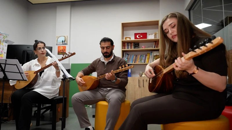 Bağlama dersi alan üç öğrenci, bir müzik okulunda birlikte çalıyor. Öğrencilerin arka planda müzik kitapları ve Türk bayrağı bulunan bir odada oturduğu görülüyor.