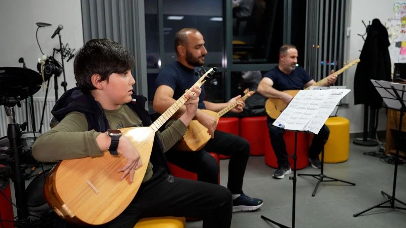 Üç kişinin bağlama çaldığı bir müzik dersi, bir öğrenci ve iki eğitmen, notaların yer aldığı bir müzik standı önünde.