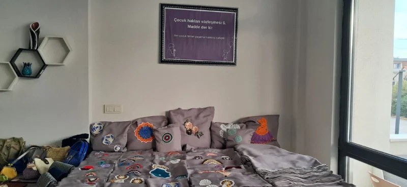 Bir odada, üzerinde el yapımı ürünlerin sergilendiği bir yatak ve duvarda çocuk hakları ile ilgili bir mesaj içeren bir tablo yer almakta.