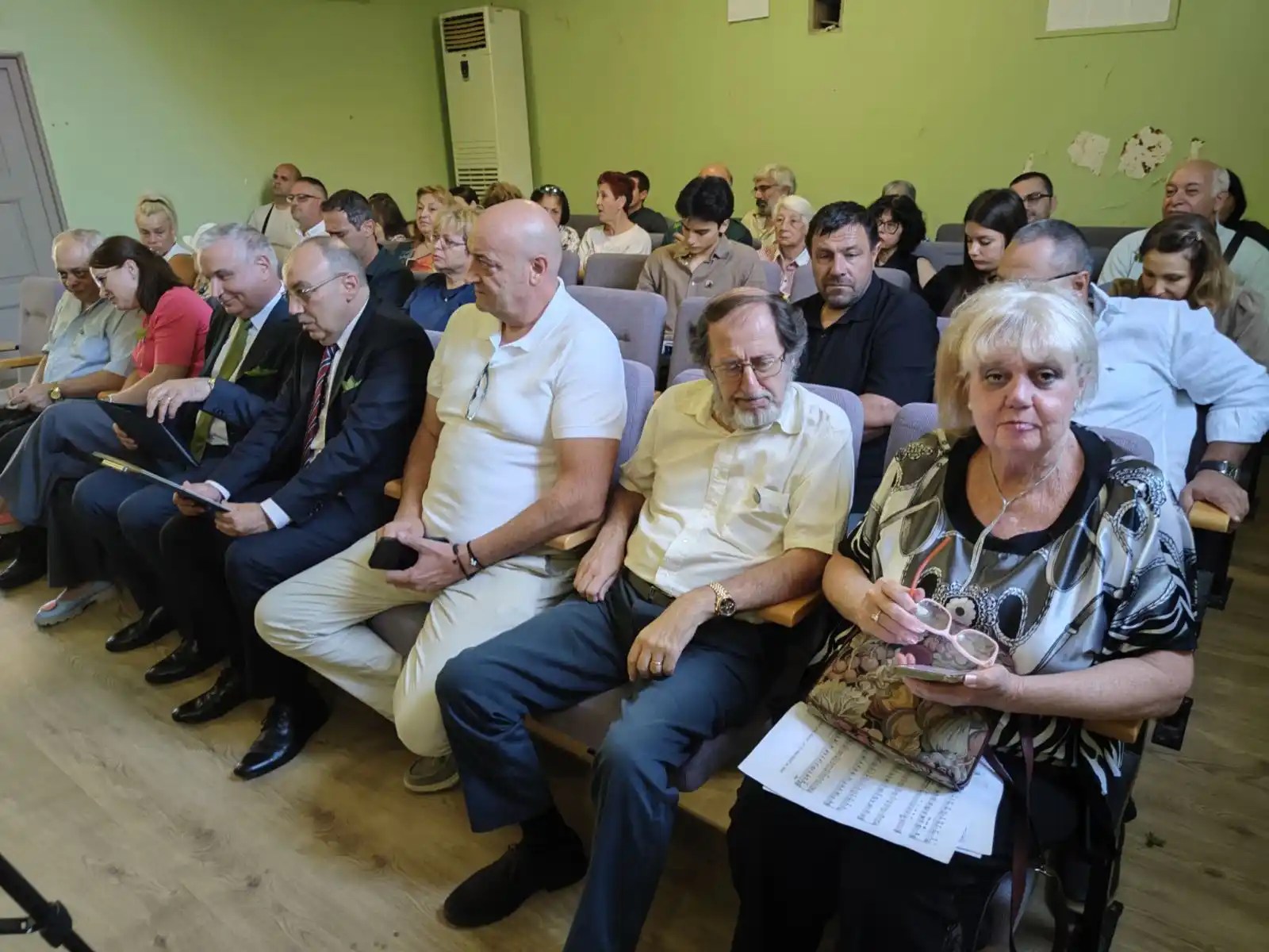 Събрание с множество участници, седящи на столове, слушащи представяне или дискусия.