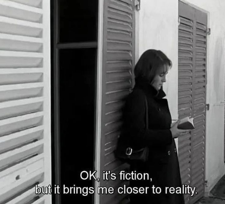 Black-and-white film still of a woman in a dark coat reading a book against white shutters, subtitled "OK, it's fiction, but it brings me closer to reality."