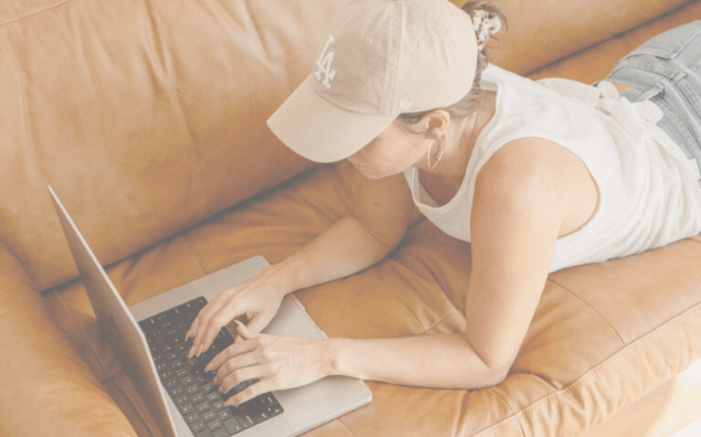 A woman in a cap and tank top lies on a tan leather couch, focused on typing on a laptop working on creating a Pinterest Business Account. The scene conveys a casual, relaxed atmosphere.