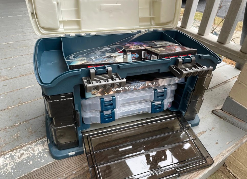 A green and white tackle box re-tasked for use as a game master toolbox. It includes a number of card decks in its top compartment. The center section includes a Savage Worlds rulebook and plastic trays.