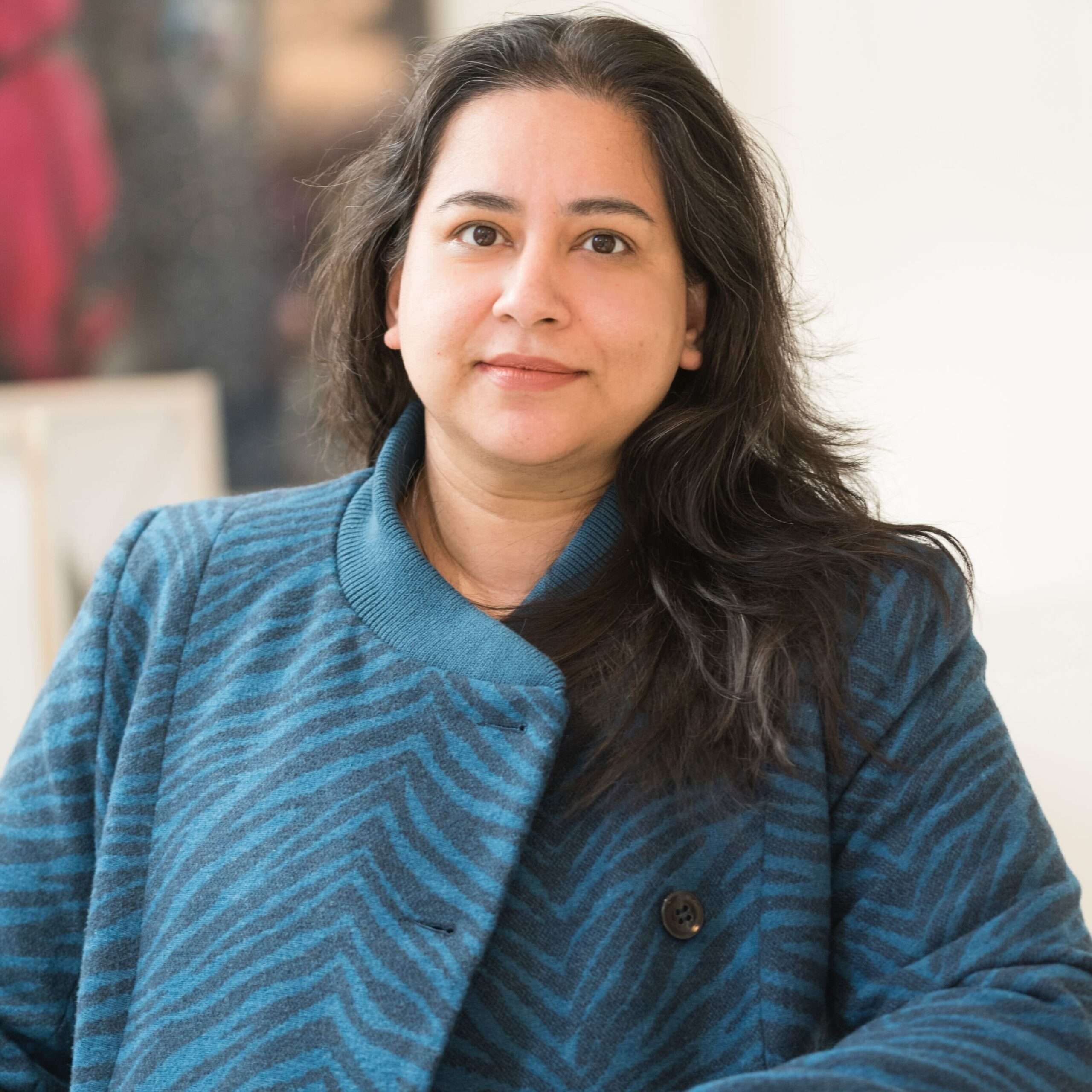 The photograph is of Naima Rashid smiling into the camera. She is dressed in a blue and black coat in a zebra pattern. Behind her is a blurry painting on a wall.