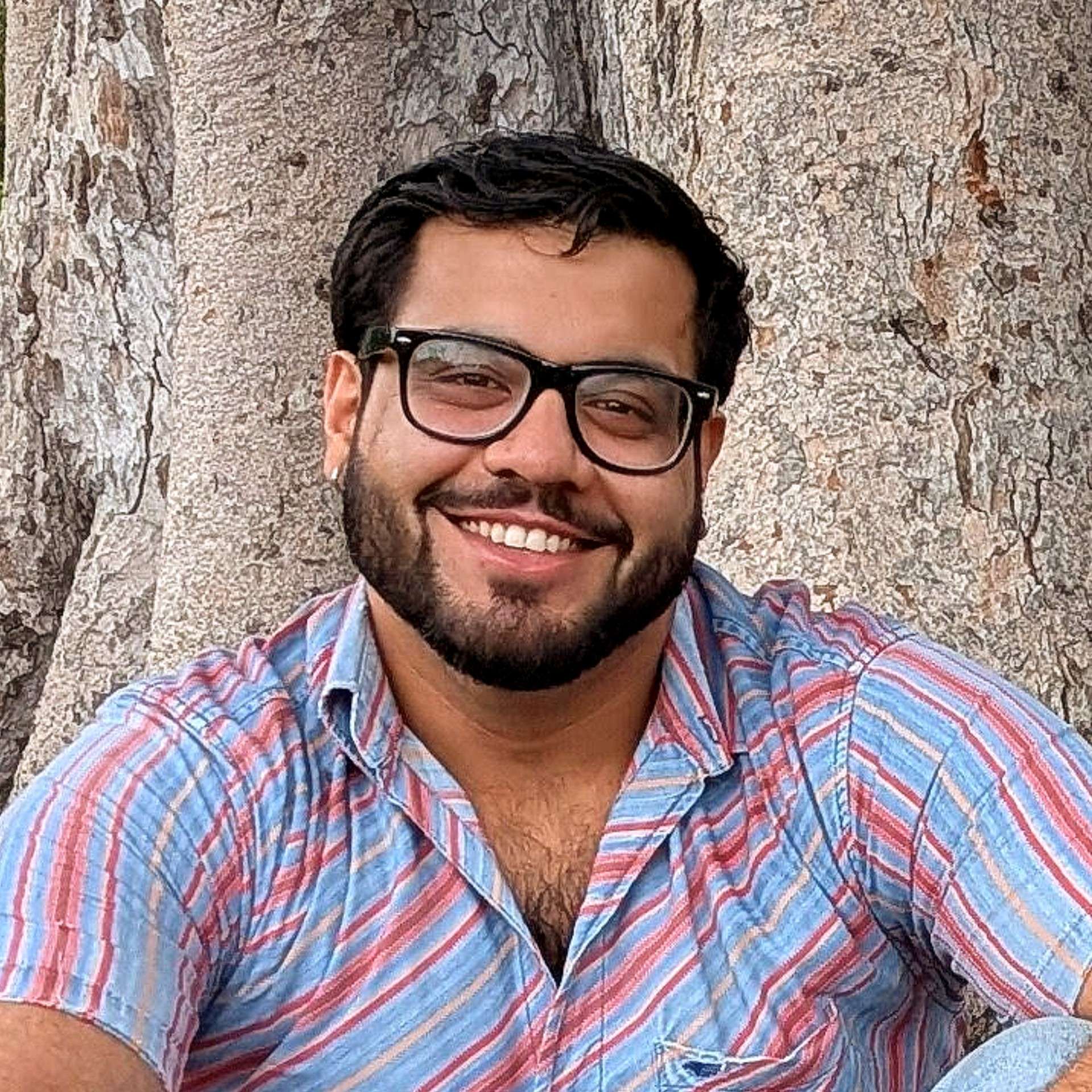 Color photo of Rishabh Pandit smiling into the camera, in daylight, in front of the trunk of a tree