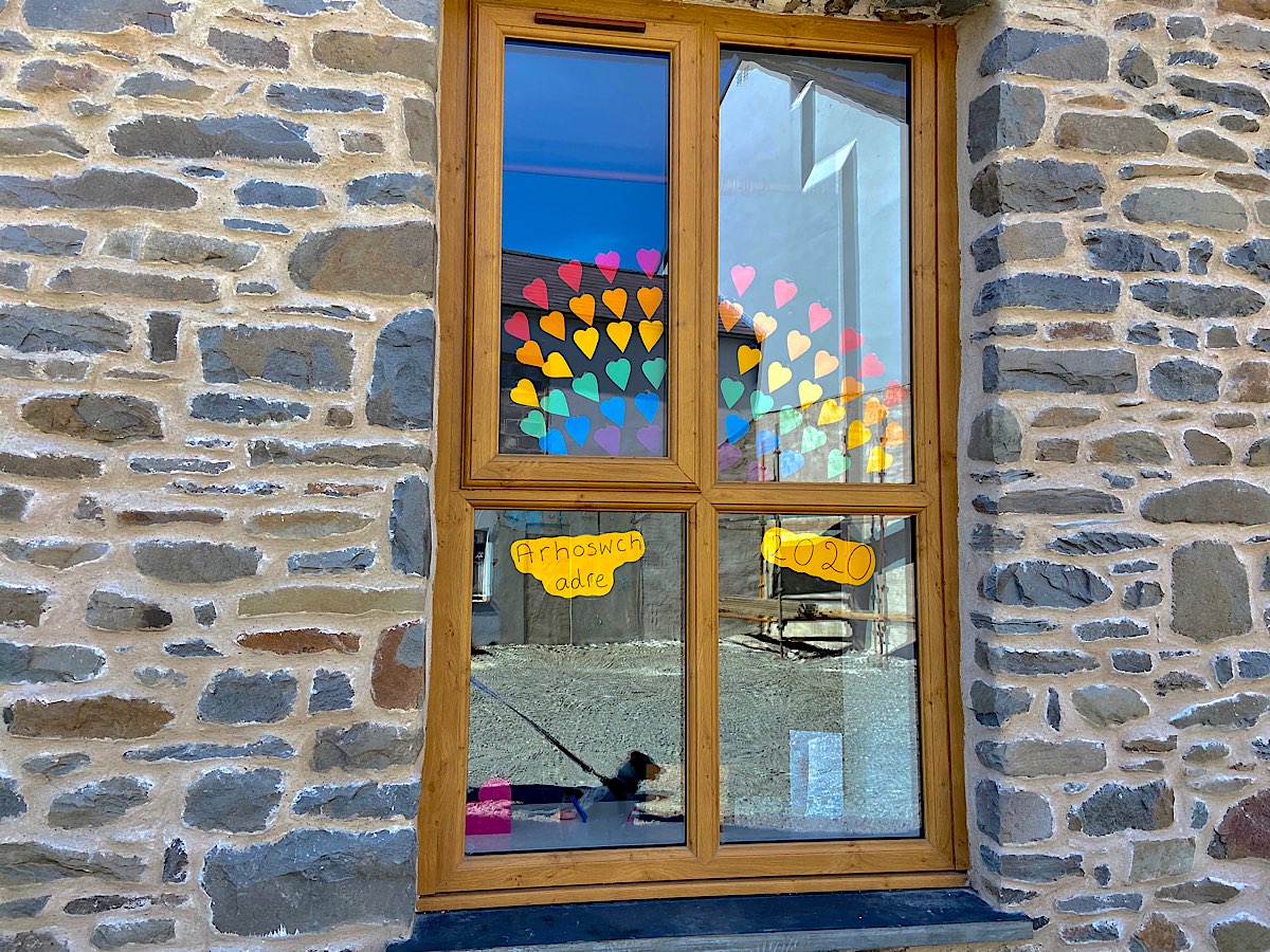 A window with a rainbow made out of coloured heart shapes. On one side it says 'Arhoswch adre' and the other side say 2020
