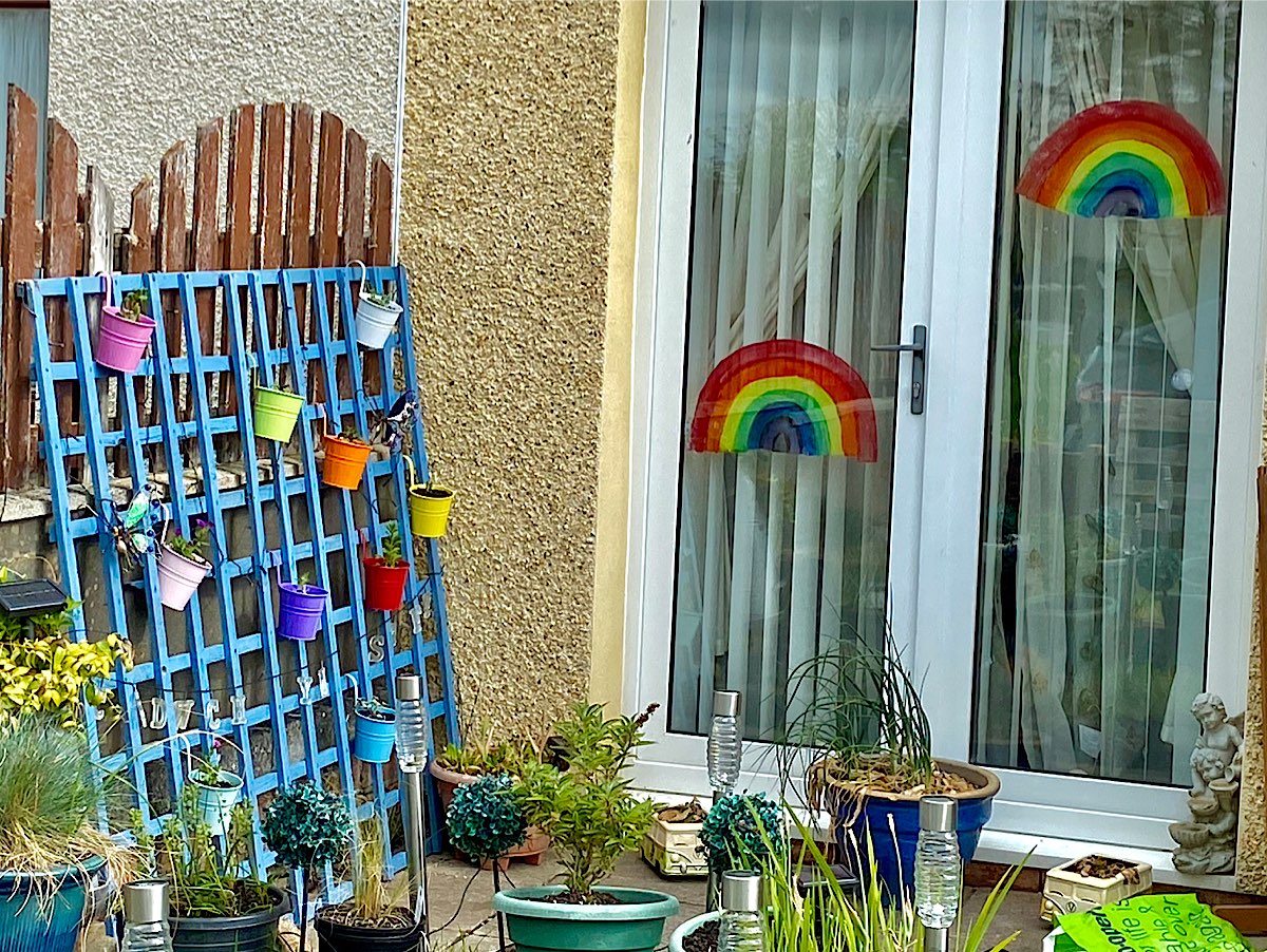 A really colourful picture with a rainbow inside a window looking out onto a trellis hung with multi-coloured plant pots