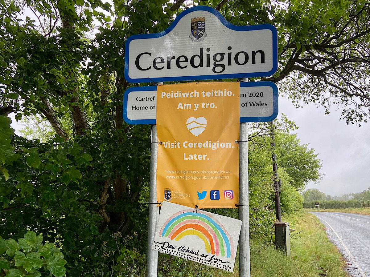 A rainbow painted on white board, says 'daw eto haul ar fryn' underneath and a small heart shaped also rainbow coloured heart sits under the rainbow. It is mounted onto the sign for Ceredigion, underneath a banner that states Ceredigion is closed