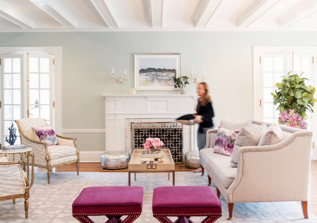 Lara Allen Brett blurred, in motion, carrying a throw pillow in a living room with a cream couch and fuschia upholstered stools.
