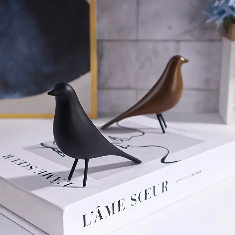 A pair of minimalist bird figurines in black and brown resin, styled on a white coffee table book with soft modern decor in the background.