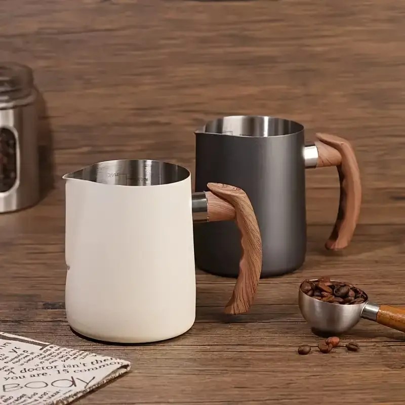Set of two coffee frothing jugs in matte white and charcoal grey, each with a stainless steel interior and sculpted wooden handle, styled on a rustic wooden surface with coffee beans and accessories.