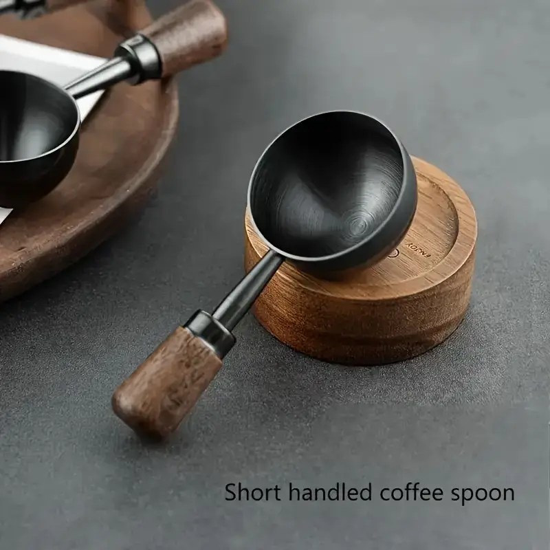 Matte black short handled coffee scoop with walnut wood handle, styled on a dark grey surface beside a wooden container.