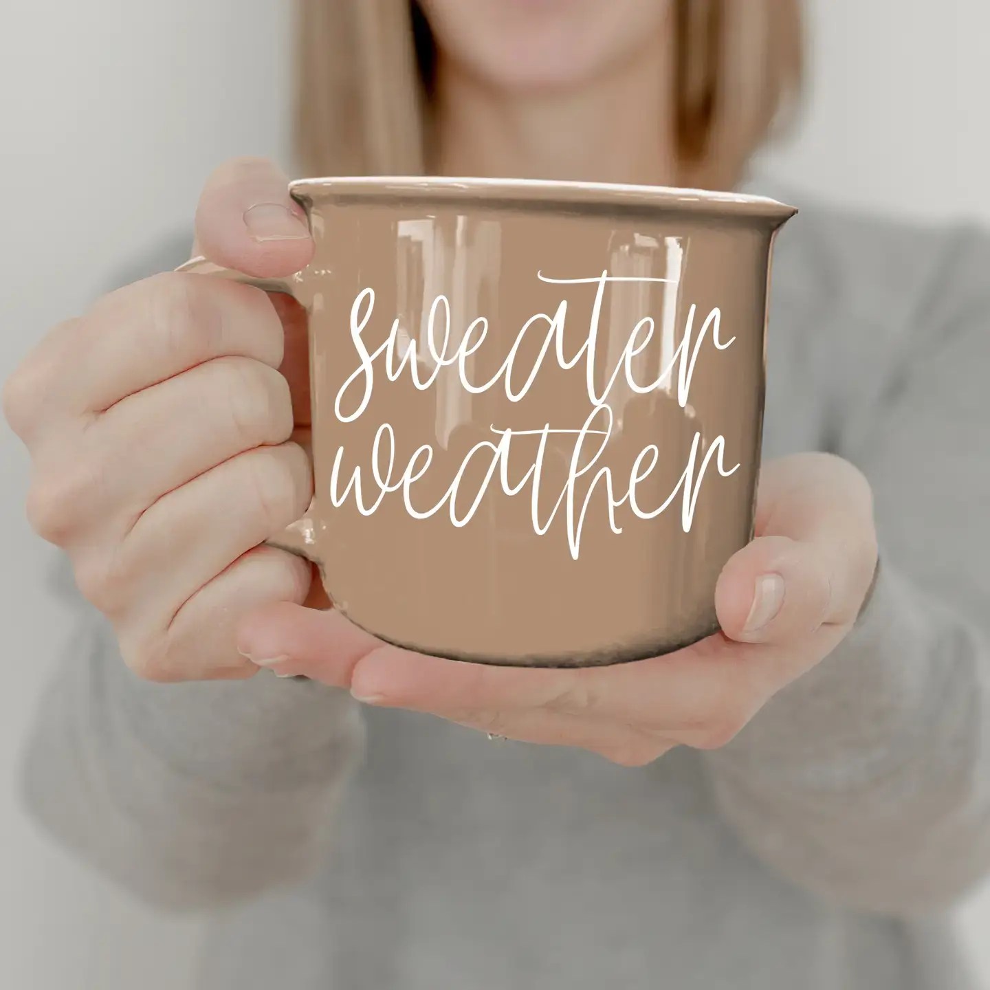 Neutral stoneware mug with “Sweater Weather” in white script, held by a person in a soft grey jumper.