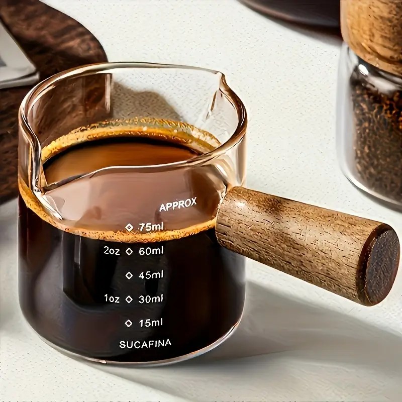 A clear glass coffee pourer with wood handle, filled with espresso, showing volume markers.