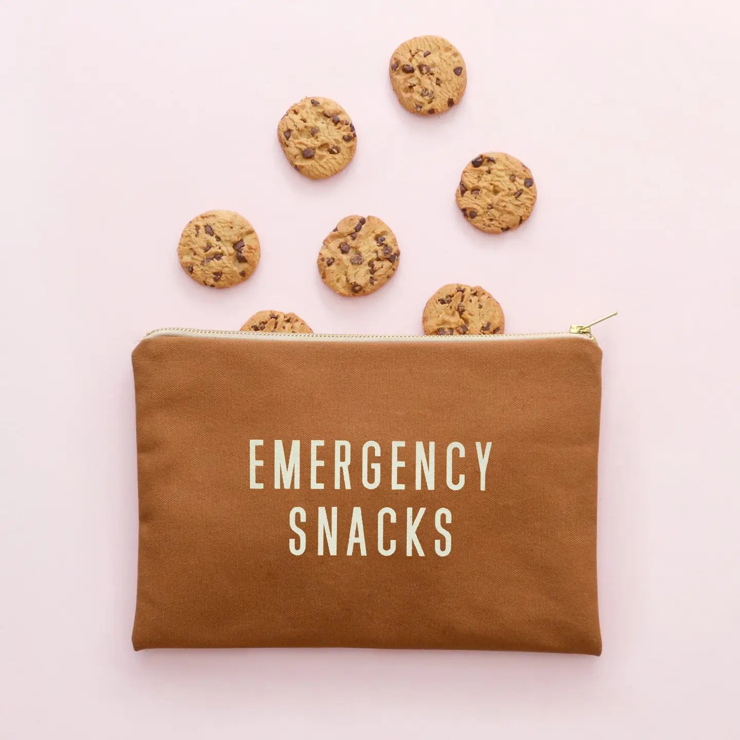 Tan "EMERGENCY SNACKS" pouch with chocolate chip cookies spilling out, set on a light pink background.