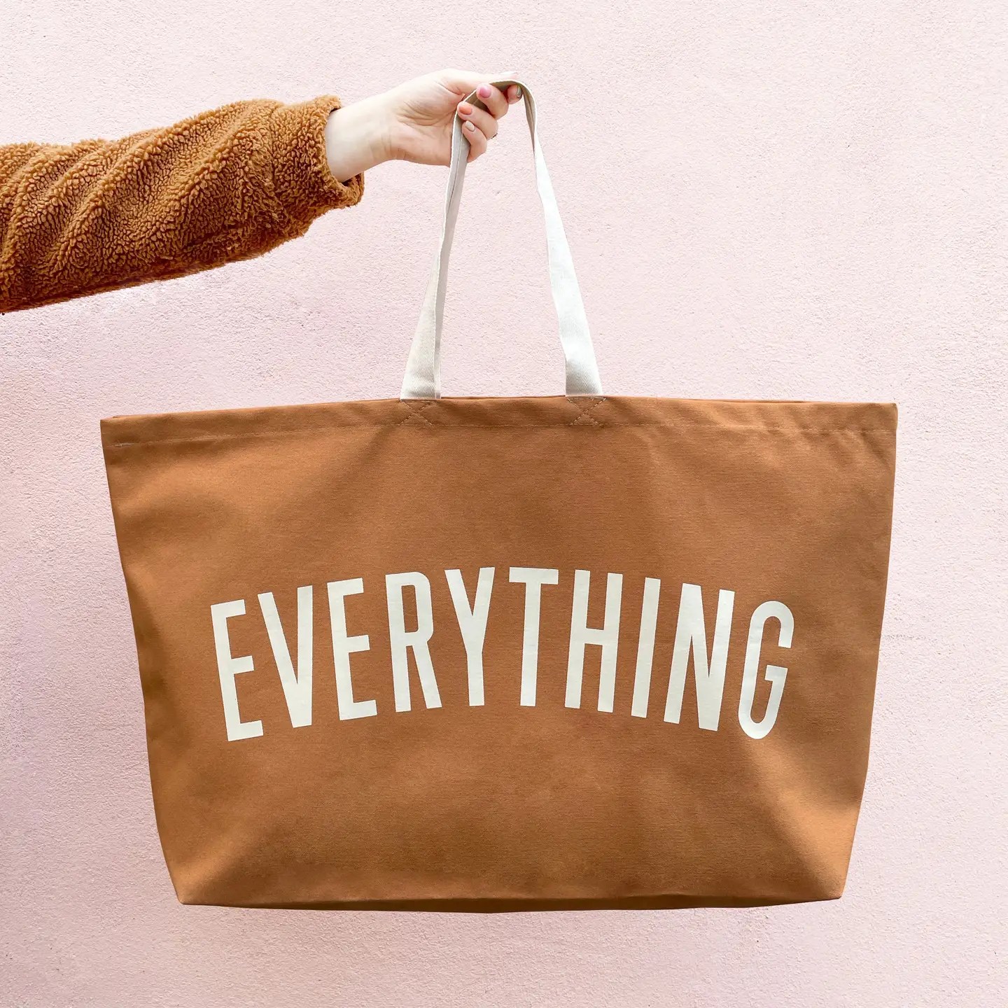 Close-up of a tan coloured "EVERYTHING" tote held by its cream strap, partially showing the printed text against a pink wall.