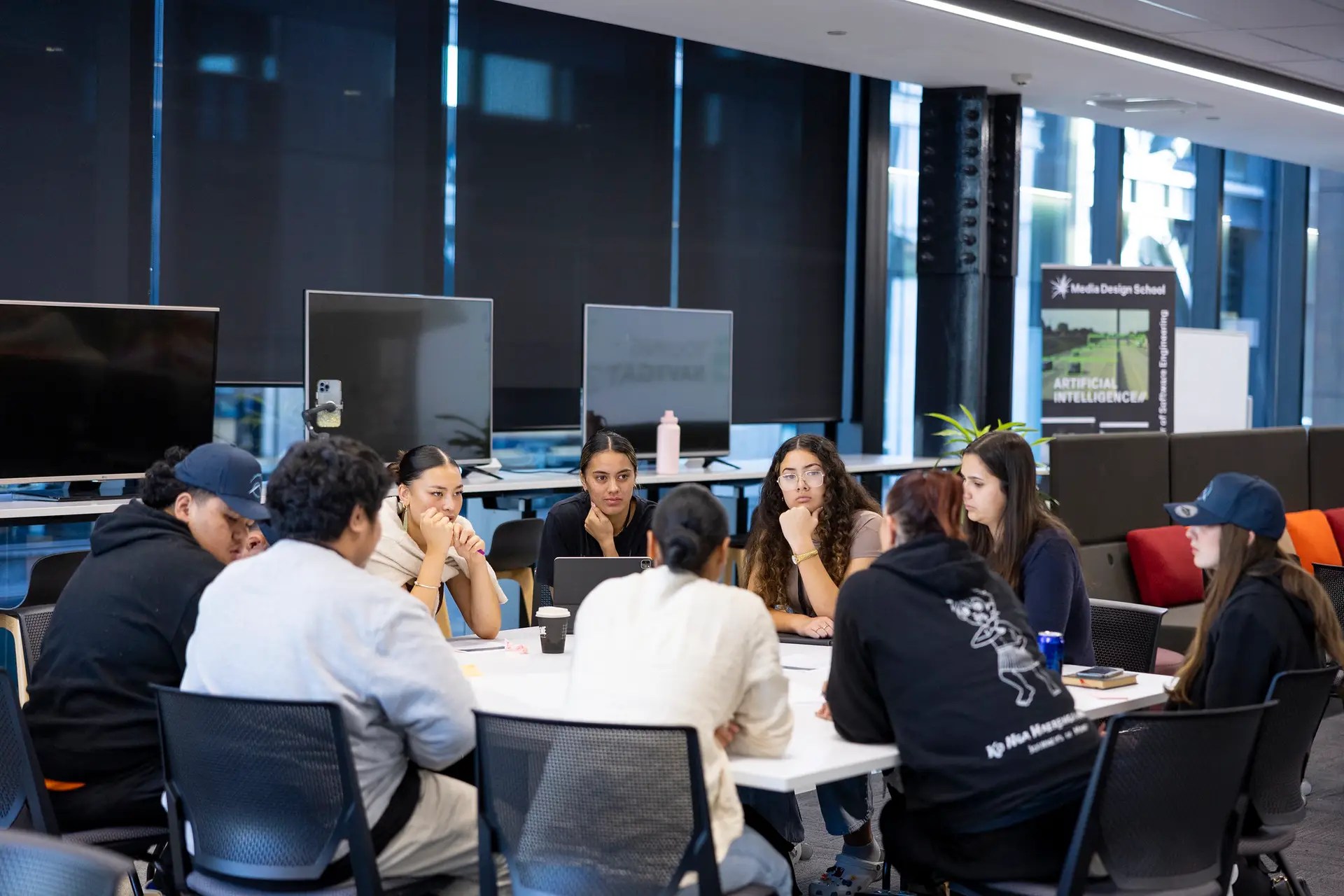 A group of young Māori and Pasifika students collaborate around a table during a startup workshop at Media Design School, discussing ideas in technology and artificial intelligence.