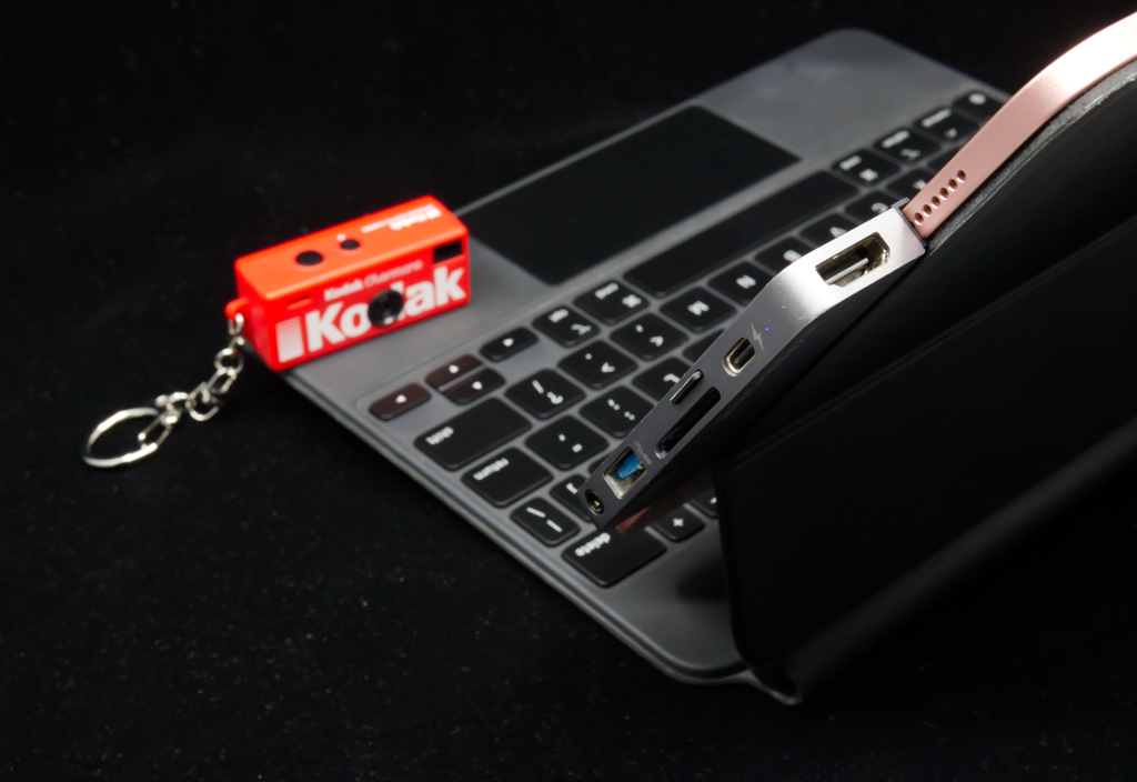 a small Kodak Charmera (a keychain digital camera) on an iPad keyboard. An iPad Air with a dongle and microSD card faces away from the camera