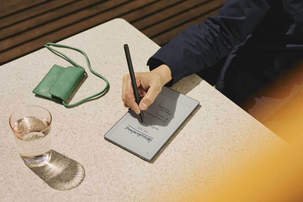 A person writes on the reMarkable Pro Paper Move tablet on a table. It's a little larger than their hand and a bit taller than a sheet of A6 paper