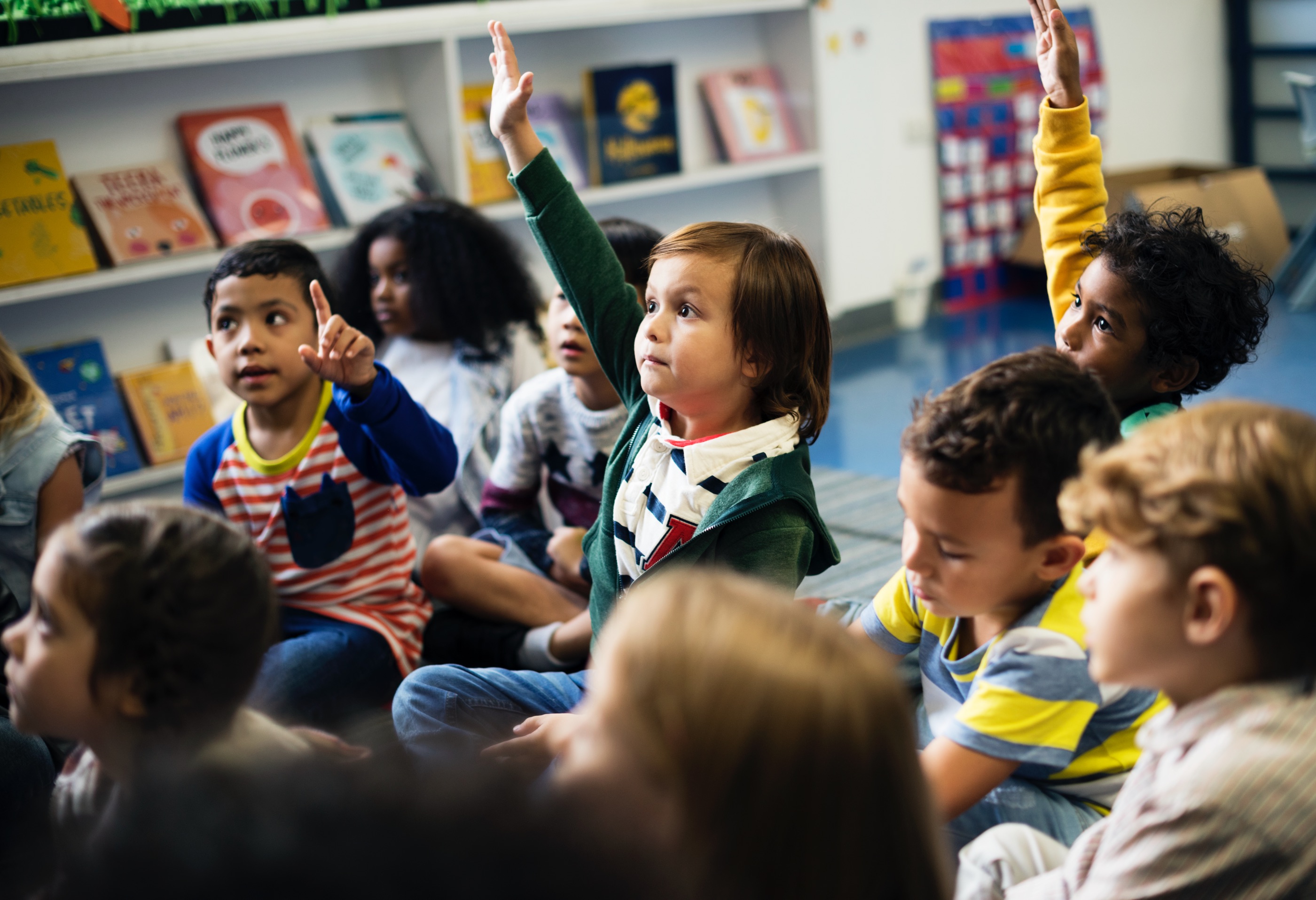 diverse group of children in the classroom raising their hands.