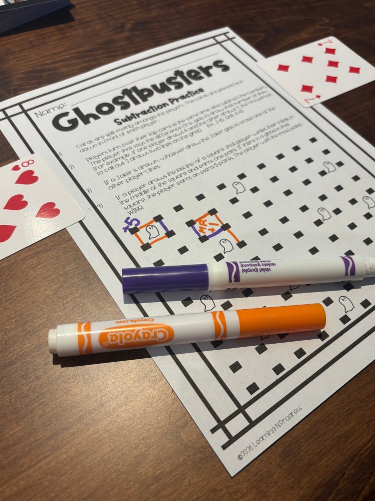 A subtraction practice worksheet titled 'Ghostbusters' with a grid, featuring cartoon ghosts, two colorful Crayola markers (orange and purple), and playing cards in the foreground.