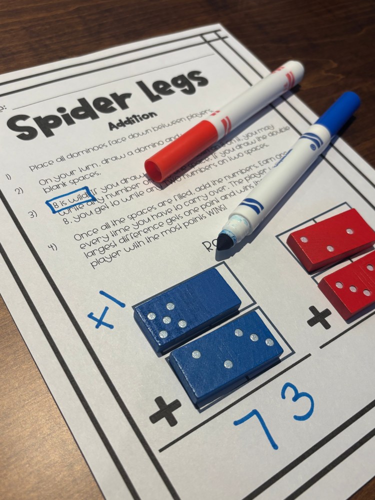A close-up of a math worksheet titled 'Spider Legs' focusing on addition, with colorful markers and dominoes on a wooden table.