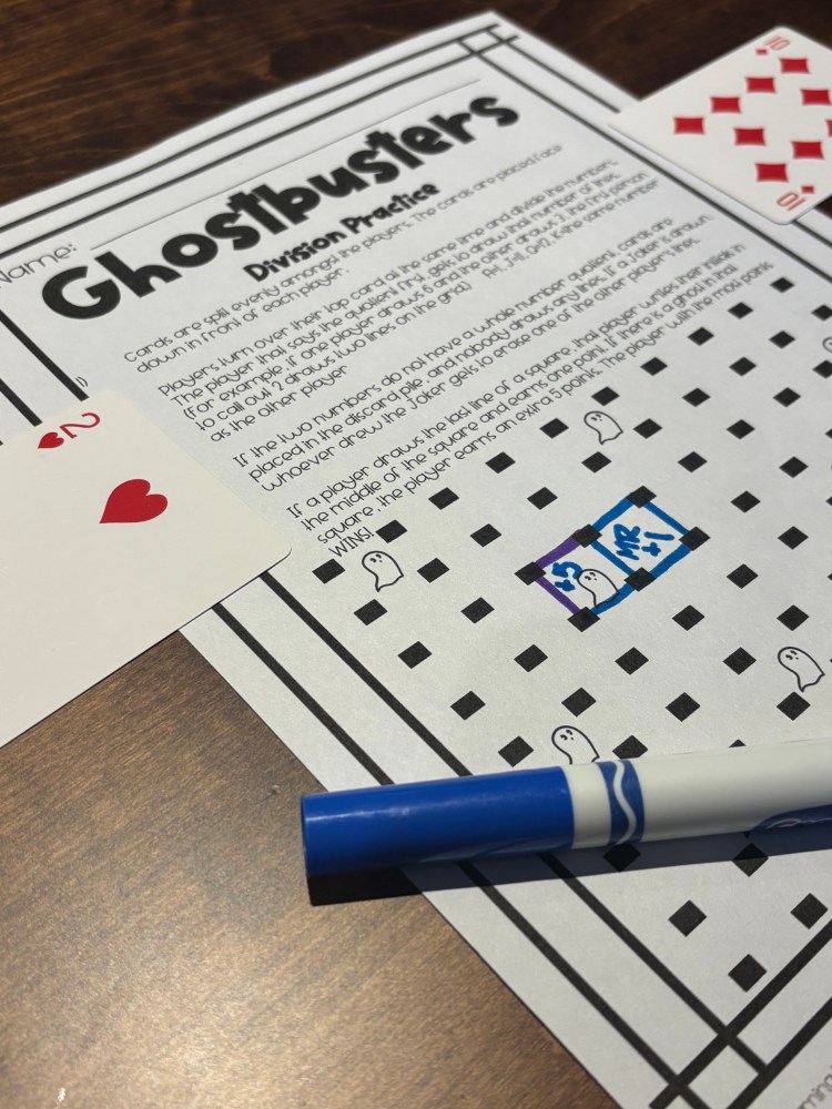 A printed worksheet titled 'Ghostbusters Division Practice' featuring a grid with playful ghost illustrations, alongside two playing cards and a blue marker on a wooden table.