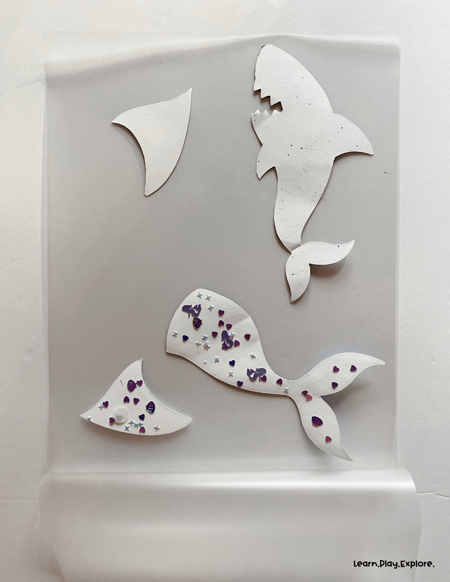 mermaid tail and shark fin printables sitting on a laminating sheet with sequence on the back. another shark fin and a shark cut out are ready to laminate as well
