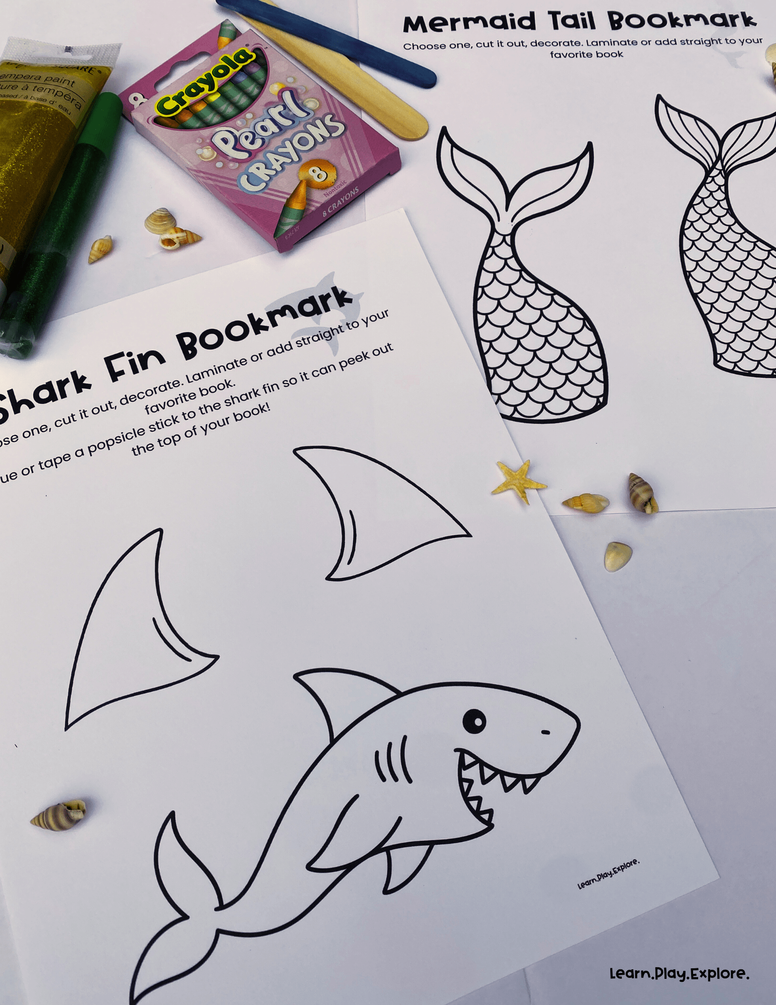 shark and mermaid printables on a table ready to be colored and cut out to turn into bookmarks there are pear crayons and glitter glue on the table