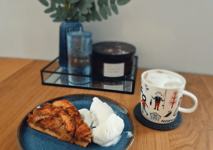 A slice of easy Norwegian eplekake served with scoops of vanilla ice cream and a frothy coffee in a Norway-themed mug, set on a wooden table with cozy decor in the background.