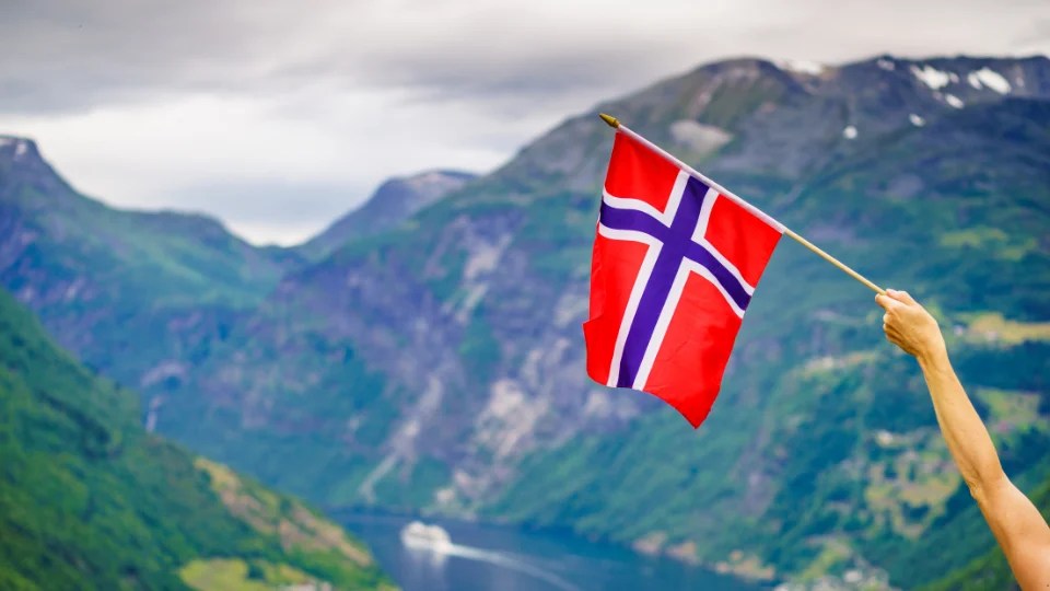 Person holding a Norwegian flag in front of a fjord and mountains — symbolizing the cultural and linguistic landscape of Norway, shaped by both Bokmål and Nynorsk.