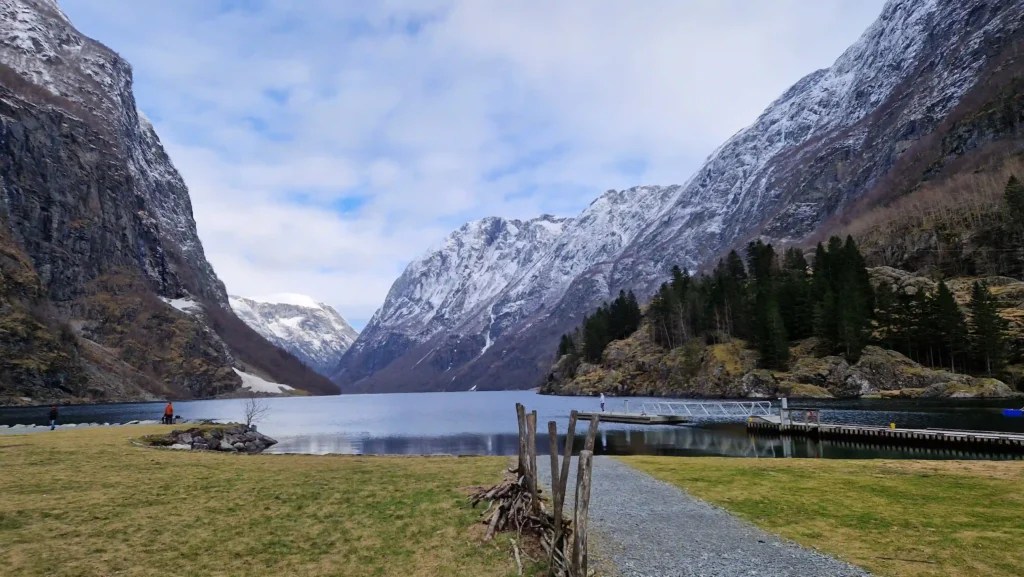 View of Gudvangen with calm fjord waters framed by steep, snow-capped mountains and grassy shores, highlighting the dramatic landscape at the end of Nærøyfjord.
