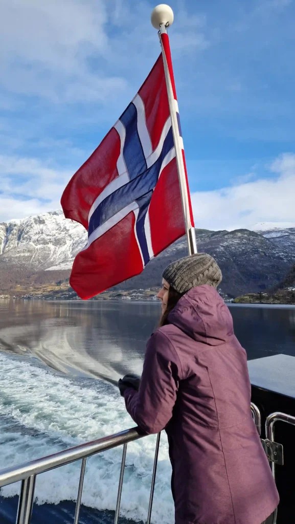 Yanitsa looking out over Aurlandsfjord from a boat, with the Norwegian flag waving above and snow-covered mountains in the background.