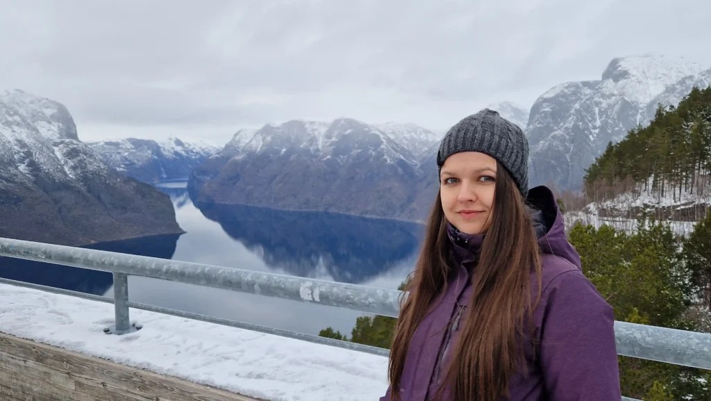 Yanitsa standing in front of a scenic view over Aurlandsfjord at Stegastein Viewpoint, with dramatic mountain peaks and fjord waters in the background.