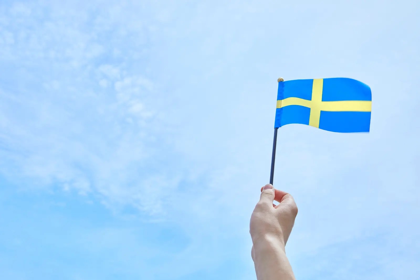 Hand holding a small Swedish flag against a blue sky - a simple symbol for starting your journey with Swedish pronunciation.