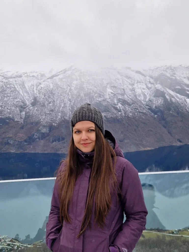 Yanitsa, the founder of Learn Scandi, standing in front of snow-covered mountains and a fjord, wearing a purple winter jacket and grey knit hat.