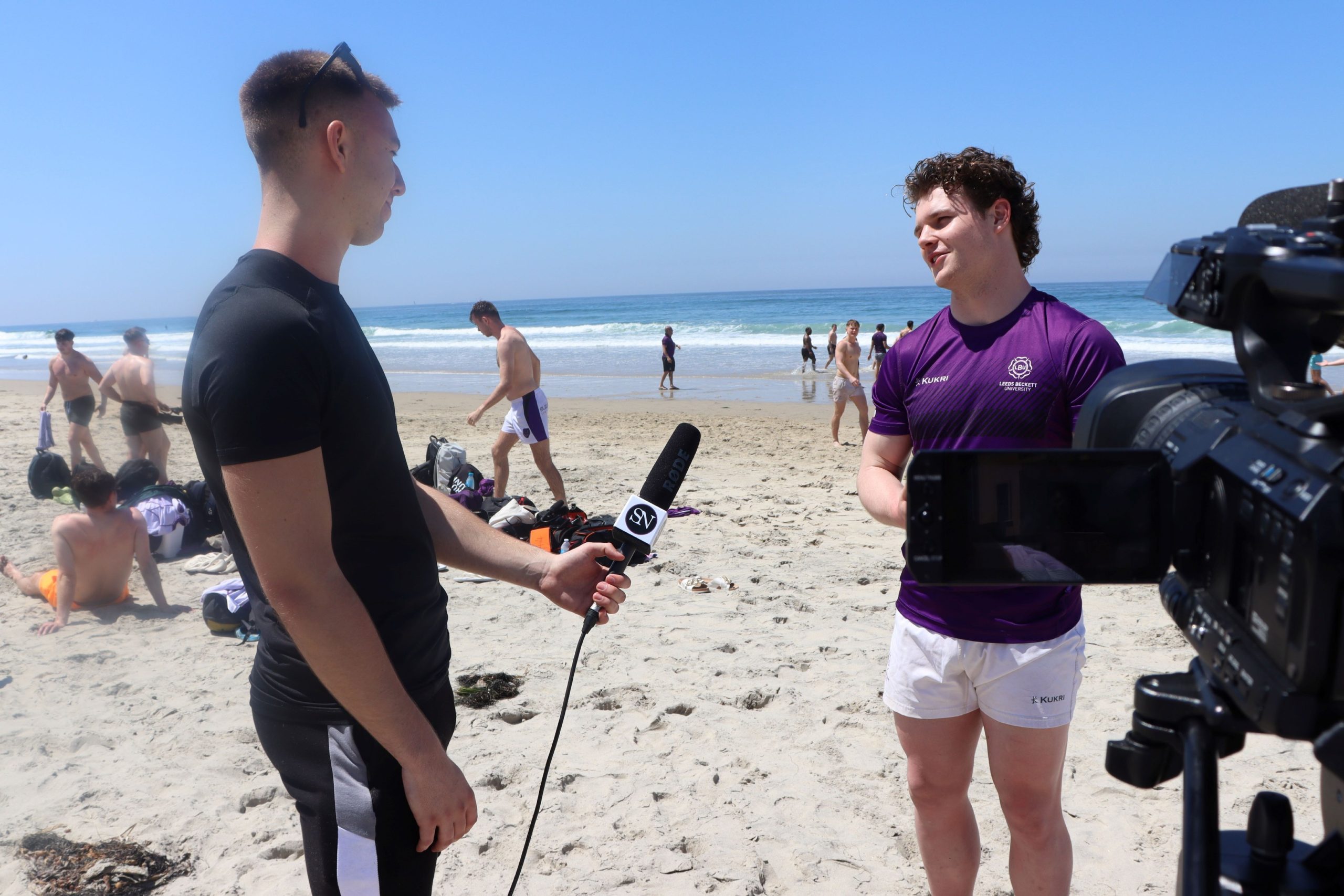 A man (Oliver Mould) interviewing another man (Alex Green) on a beach