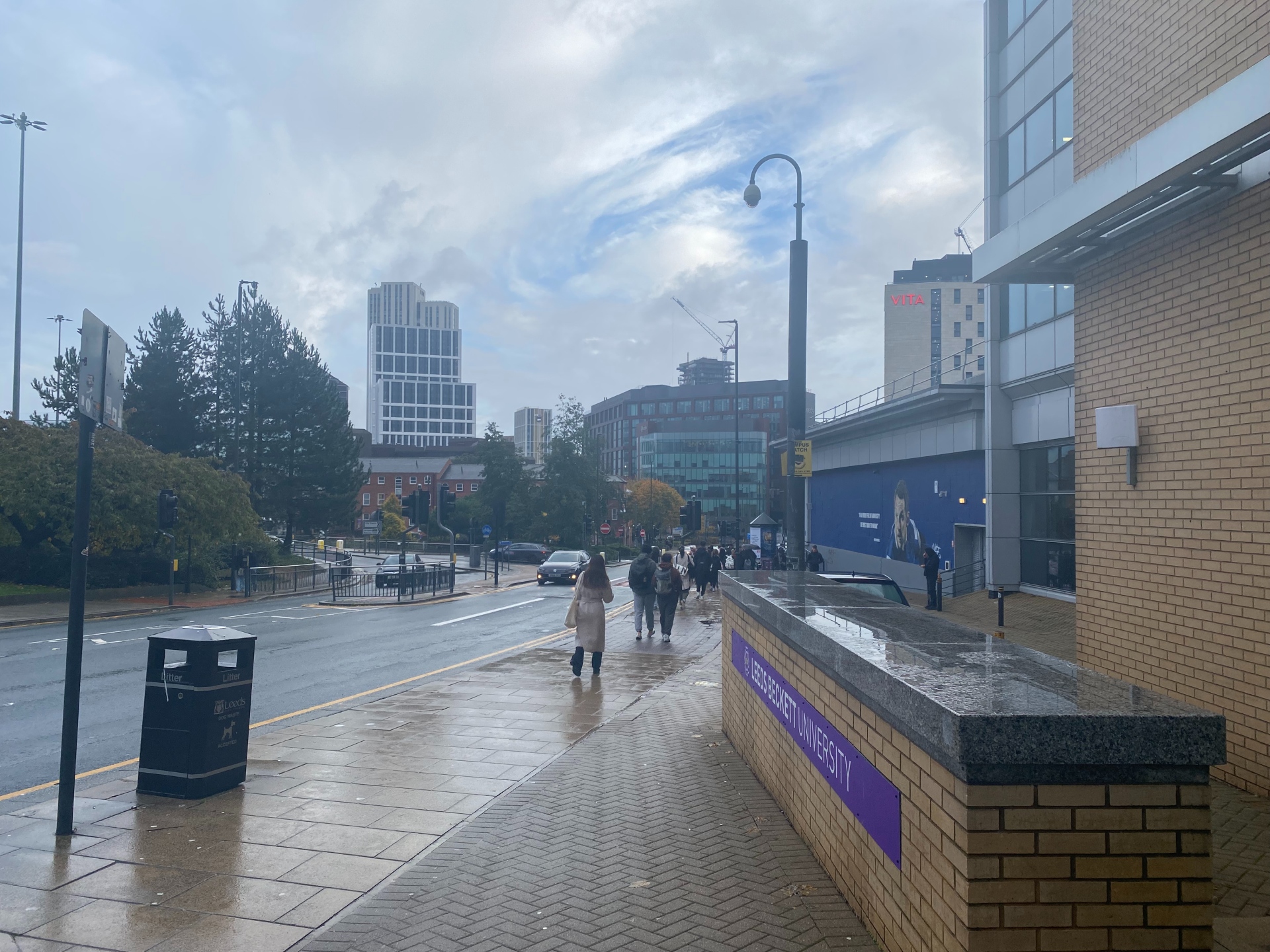 The image shows a Leeds Becket University sign with standing water on top. The city of Leeds is in the background with high rise buildings and people walking in the street.